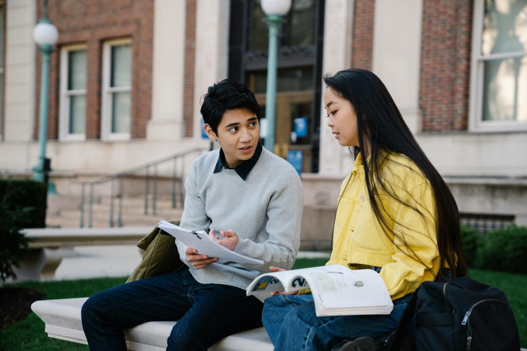Two students discussing notes on a campus bench, surrounded by academic buildings.